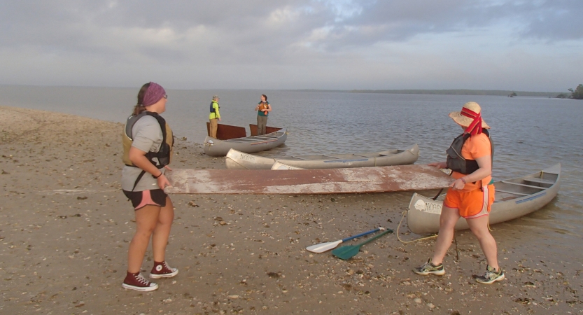 In the foreground, two people carry what appears to be a panel of wood across a beach. Behind them, canoes rest on the shore.